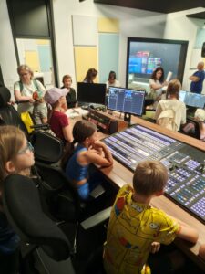 Skupina detí a dospelých sedí v televíznej réžii pri veľkom mixážnom pulte a monitory, kým im inštruktorka v pozadí niečo vysvetľuje.