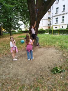 Tri dievčatá sa hrajú vonku v parku na trávnatej ploche, pričom jedno dievča šplhá po lane uviazanom na strome.
